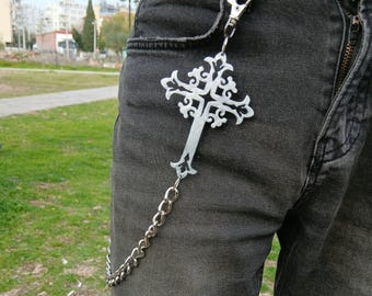 May include: A silver chain with a decorative cross pendant attached to it. The chain is hanging from a person's belt loop.