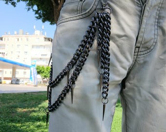 May include: Gunmetal-colored chain accessory with multiple strands and spiked accents. The chains are attached to a clasp and hang down, creating a layered effect. The accessory is shown attached to a pair of light-colored jeans.