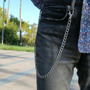 May include: A silver-colored chain attached to a pair of dark gray denim jeans. The chain is made of interlocking oval links and is clipped to the belt loop and pocket. The jeans have a classic five-pocket design.