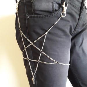 May include: A silver chain body harness with a star-shaped design and a small pendant hanging from the bottom. The harness is worn over black jeans.