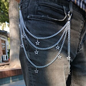 May include: Silver chain belt with multiple strands and star charms. The belt is attached to the jeans with a clasp. The chains hang down, creating a layered effect. The belt is a fashion accessory.