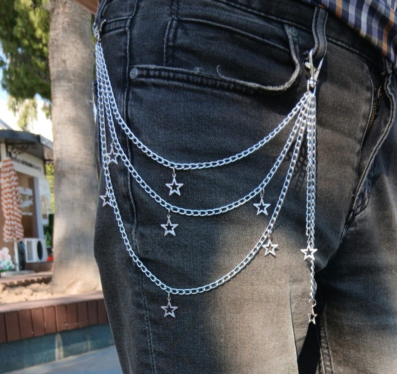 May include: Silver chain belt with multiple strands and star charms. The belt is attached to the jeans with a clasp. The chains hang down, creating a layered effect. The belt is a fashion accessory.