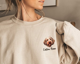 May include: A beige sweatshirt with a small pocket on the left side. The pocket has a brown and white dog's face printed on it. The text "Custom Name" is printed below the dog's face.