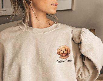 May include: A beige sweatshirt with a small, embroidered image of a brown dog with a happy expression. The text "Custom Name" is printed below the dog.