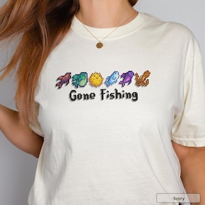 May include: Ivory-colored t-shirt with a pixelated fishing-themed graphic. The design includes various colorful fish and the text "Gone Fishing" in black. The shirt is a casual, short-sleeved style.