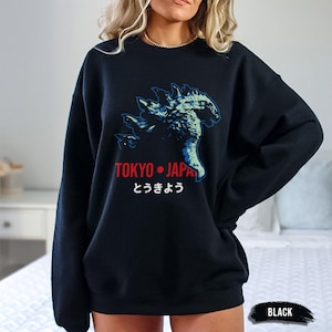 May include: A black crewneck sweatshirt featuring a graphic of a blue and green monster silhouette. Below the graphic, the text reads TOKYO JAPAN in red, followed by the Japanese characters とうきょう in white. The sweatshirt has long sleeves.