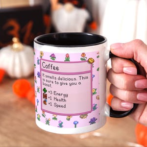 May include: White coffee mug with a black interior and handle. The mug features a pink label with the word "Coffee" and text that reads "It smells delicious. This is sure to give you a boost." with pixelated graphics.