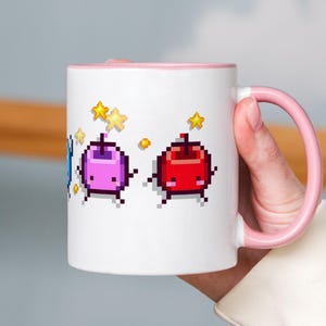 Könnte beinhalten: Eine weiße Keramiktasse mit rosafarbenem Rand und Henkel. Die Tasse zeigt ein Pixel-Design mit vier Figuren, von denen drei rot, lila und blau sind. Die Figuren sind von gelben Sternen umgeben.