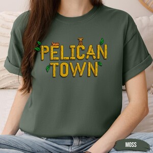 May include: A moss green t-shirt with the text "PELICAN TOWN" in yellow letters with a small graphic of a dog and a leaf design.