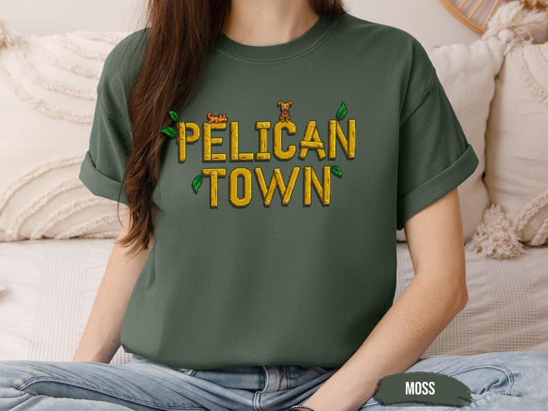 May include: A moss green t-shirt with the text "PELICAN TOWN" in yellow letters with a small graphic of a dog and a leaf design.