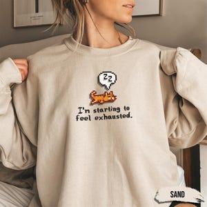 May include: A beige sweatshirt with a pixelated image of a dog sleeping with a speech bubble above it that says "zzZ". The text below the image says "I'm starting to feel exhausted."
