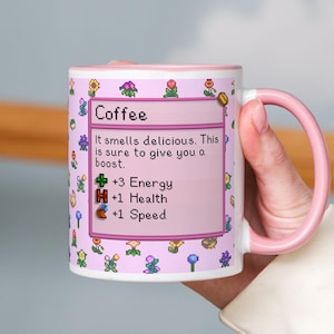 Könnte beinhalten: Eine weiße Keramik-Kaffeetasse mit rosa Innenraum und Griff. Die Tasse hat einen rosa Hintergrund mit pixeligen Blumen und dem Text "Coffee". Der Text auf der Tasse lautet "It smells delicious. This is sure to give you a boost. +3 Energy, +1 Health, +1 Speed."