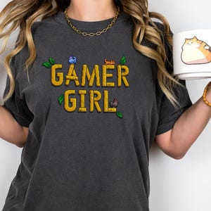 May include: A dark gray t-shirt with the text "GAMER GIRL" in yellow pixelated letters. The shirt has a small green leaf design and pixelated images of a blue bird, a red cat, and a brown chicken.