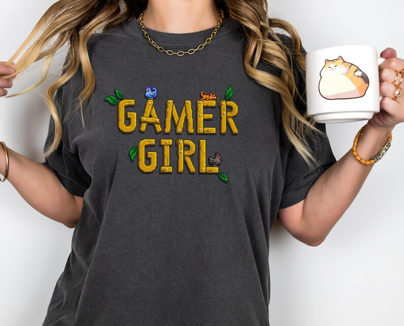 May include: A dark gray t-shirt with the text "GAMER GIRL" in yellow pixelated letters. The shirt has a small green leaf design and pixelated images of a blue bird, a red cat, and a brown chicken.