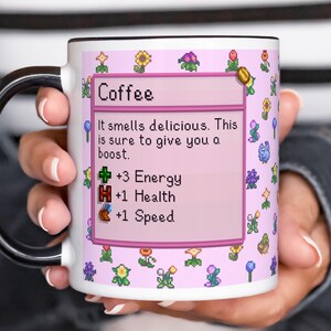 May include: White ceramic coffee mug with a black handle and rim. The mug features a pink background with pixelated flowers and a text box that reads "Coffee". The text box also includes the phrases "It smells delicious. This is sure to give you a boost."