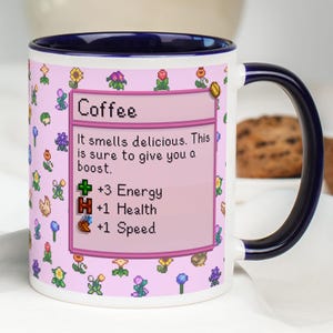 May include: White ceramic coffee mug with a dark blue handle and rim. The mug features a pink background with pixelated flowers and a text box that reads "Coffee". The text box also includes the phrases "+3 Energy", "+1 Health", and "+1 Speed".