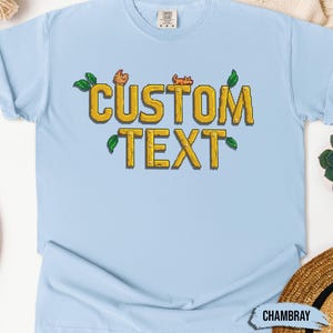 May include: Light blue t-shirt with a pixelated design of the words "CUSTOM TEXT" in yellow with green leaves and small orange pixelated animals.
