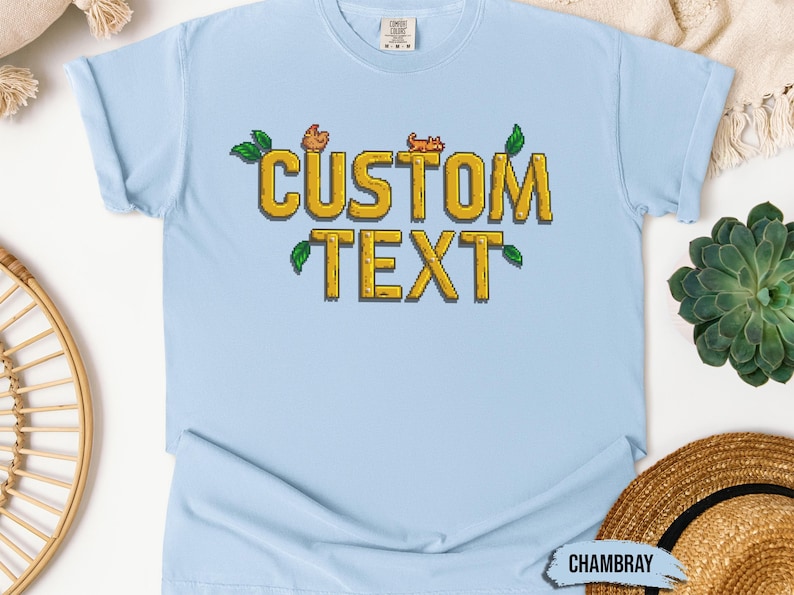 May include: Light blue t-shirt with a pixelated design of the words "CUSTOM TEXT" in yellow with green leaves and small orange pixelated animals.