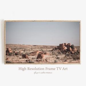 May include: A framed photograph of a desert landscape with large, rounded red rocks in the foreground. The sky is a pale blue and the ground is covered in dry, brown grass.