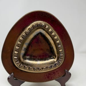 May include: A triangular, vintage ashtray with a dark red and gold color scheme. The ashtray has a textured, crackled finish and a raised, gold-colored inner section. It is displayed on a small, dark brown wooden stand.