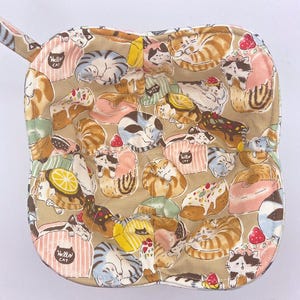 May include: Tan quilted oven mitt with a loop for hanging. The mitt is patterned with various sleeping cats, desserts, and the text "Hello Cat". The design includes pink, brown, and blue elements.