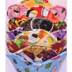 May include: A stack of colorful bowl cozies with various food-themed patterns. The patterns include sushi, coffee beans, grapes, and pizza. The cozies are made of fabric and are designed to insulate bowls.