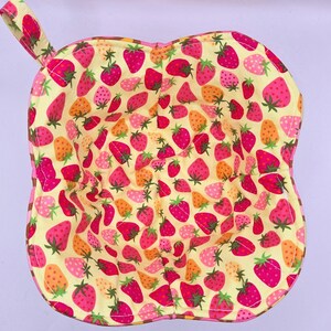 May include: A handmade bowl cozy with a strawberry pattern. The cozy is yellow with a repeating pattern of pink and orange strawberries with green stems. The edges are pink and brown, and it has a small handle.