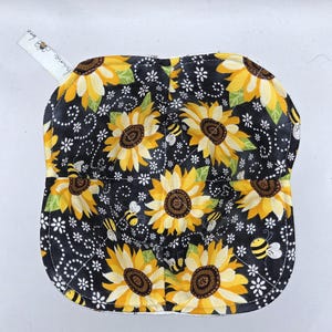 May include: A square bowl cozy with a black background, featuring yellow sunflowers and bees. The cozy has a white trim and a small white tag with text and a bee graphic. The sunflowers have brown centers and green leaves.