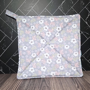 May include: Square gray pot holder with a floral pattern featuring white and pink flowers, and light green leaves. The pot holder has a loop for hanging and is quilted with diagonal stitching.