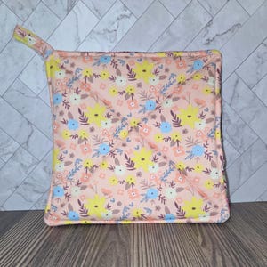 May include: A square, quilted pot holder with a floral pattern. The fabric features a light pink background with yellow, blue, and peach-colored flowers and green leaves. A yellow loop is attached for hanging.