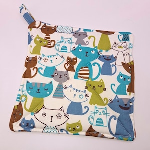 May include: A white quilted hot pad with a pattern of colorful cartoon cats. The cats are in various poses and colors, including blue, green, brown, and gray.