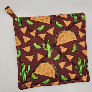 May include: A brown quilted hot pad with a pattern of green cacti, yellow tacos, and lime wedges. The hot pad has a loop handle.