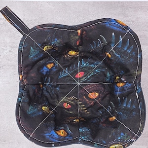 May include: A black, square-shaped pot holder with a loop for hanging. The fabric is patterned with multiple dragon eyes in shades of yellow, red, and blue. White stitching outlines the edges and creates a grid pattern across the surface.