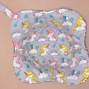 May include: A gray, quilted pot holder with a handle, featuring a repeating pattern of unicorns, rainbows, clouds, and stars. The unicorns have yellow and pink manes and horns. The pot holder has a light pink border.