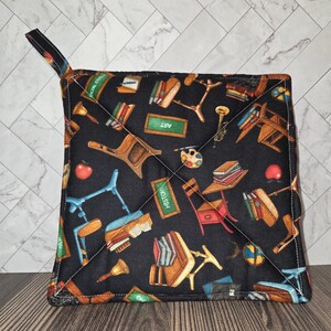 May include: Black square pot holder with a loop for hanging. The fabric features a repeating pattern of school-related items like chairs, books, apples, and the word "ART" on a chalkboard. White stitching outlines the square and the diagonal quilting.