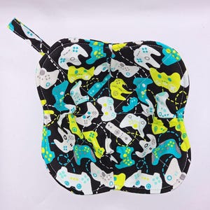 May include: Black, quilted pot holder with a loop for hanging. The pot holder is patterned with a repeating design of video game controllers in teal, lime green, and white. The edges are trimmed in lime green.