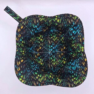 May include: A square, quilted pot holder with a loop for hanging. The fabric features a pattern of overlapping scales in shades of blue, green, and yellow on a black background. White stitching outlines the shape.