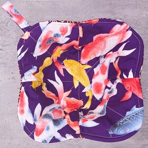 May include: A quilted, purple pot holder with a loop for hanging. The pot holder features a repeating pattern of colorful koi fish in shades of red, yellow, white, and blue. The edges are stitched with white thread.