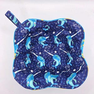 May include: A square, quilted bowl cozy with a navy blue background featuring light blue narwhals, white stars, and swirls. The cozy has a light blue trim and a small loop handle. The interior is yellow.