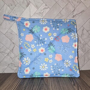 May include: Square, light blue pot holder with a floral pattern. The pattern includes white, pink, and teal flowers with green leaves. It has a small loop for hanging and is quilted with a diamond pattern.