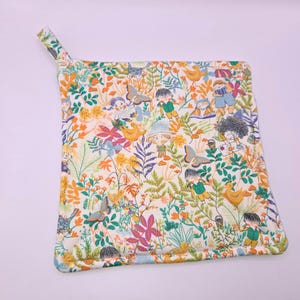 May include: A white cotton pot holder with a colorful pattern of children, birds, butterflies, and flowers. The pot holder has a loop for hanging.
