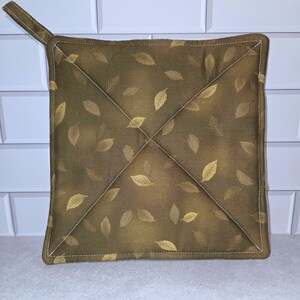 May include: Square, olive green pot holder with a loop for hanging. The pot holder features a repeating pattern of gold-colored leaves. White stitching creates a cross design in the center and outlines the edges.