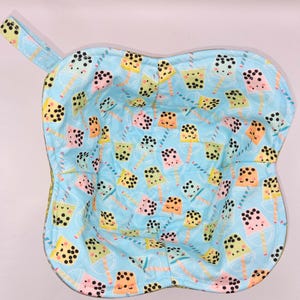 May include: A light blue fabric bowl cozy with a handle, patterned with colorful bubble tea drinks. The cozy is designed to hold a bowl and keep food warm. The bubble tea drinks are in yellow, pink, orange, and green.