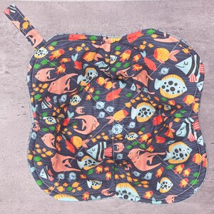 May include: A quilted, navy blue pot holder with a loop, featuring a colorful fish pattern. The design includes various fish in shades of orange, yellow, and blue, set against a wavy background. The pot holder is square with rounded edges.