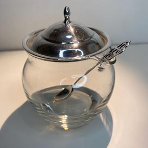 Könnte beinhalten: Eine klare Glasschüssel für Zucker mit einem silbernen Deckel und einem passenden Löffel. Die Schüssel hat eine abgerundete Form und einen strukturierten Boden.