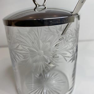Könnte beinhalten: Ein klares Glasgefäß mit einem silbernen Deckel und einem silbernen Löffel. Das Gefäß ist mit einem floralen Muster graviert. Der Deckel hat einen Ringgriff und die Worte "Sterling 920" sind in den Deckel graviert.