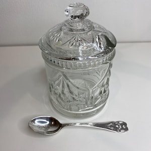 Peut inclure: Un sucrier en verre transparent avec un couvercle et une petite cuillère en argent. Le bol est décoré d'un motif de feuilles et de fleurs.