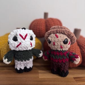 Puede incluir: Dos muñecos de ganchillo hechos a mano, uno parecido a Jason Voorhees con una máscara blanca y detalles rojos, el otro a Freddy Krueger con sombrero marrón y suéter a rayas. Detrás, calabazas de ganchillo naranjas y amarillas, perfectas para Halloween.