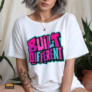May include: White t-shirt with the text "BUILT DIFFERENT" in a vibrant, graffiti-style design. The text features shades of pink, purple, and blue, outlined in black. The shirt is a casual, oversized fit.