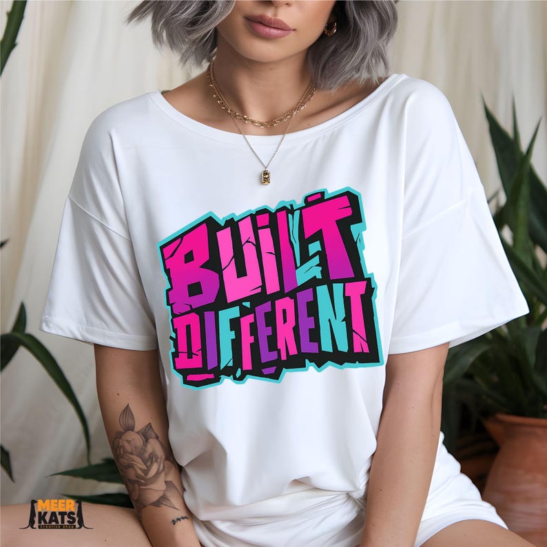 May include: White t-shirt with the text "BUILT DIFFERENT" in a vibrant, graffiti-style design. The text features shades of pink, purple, and blue, outlined in black. The shirt is a casual, oversized fit.
